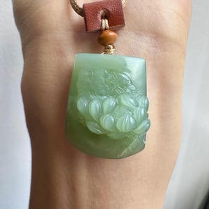 May include: A light green jade pendant featuring a detailed relief carving of a blooming flower. The pendant is suspended from a tan cord with a brown leather accent piece and a small wooden bead, creating a natural and earthy aesthetic.