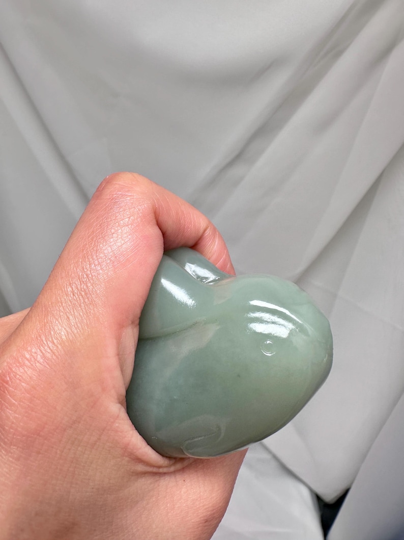 Puede incluir: Una talla de piedra verde p&aacute;lido de una ballena. La ballena se sostiene en una mano y la piedra tiene un acabado brillante. La talla tiene un dise&ntilde;o simple y redondeado con detalles sutiles.