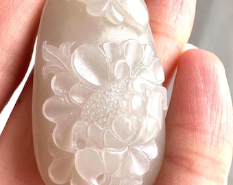 May include: A smooth, translucent, off-white stone pendant featuring a detailed relief carving of a large blooming flower with layered petals and leaves. A small pink bead is attached to the top of the pendant near the silver-toned bail.