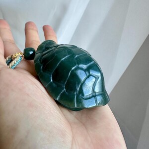 May include: A dark green carved stone turtle pendant featuring a detailed shell pattern. The turtle is attached to a decorative cord with a small matching green bead and a braided accent in shades of blue, white, and gold metallic thread.