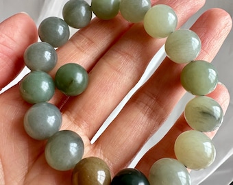 Puede incluir: Un brazalete de cuentas redondas grandes en tonos verdes, grises y marrones. El brazalete se sostiene en una mano, mostrando los diversos colores y texturas de las piedras pulidas. Las cuentas tienen aproximadamente 2,5 cm de diámetro.