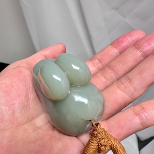Puede incluir: Un colgante de jade verde p&aacute;lido, liso y de forma org&aacute;nica. Presenta tres protuberancias ovaladas y una base redondeada. Una cuerda trenzada marr&oacute;n est&aacute; unida. El colgante se sostiene en una mano. El colgante de jade mide unos 6 cm de largo.