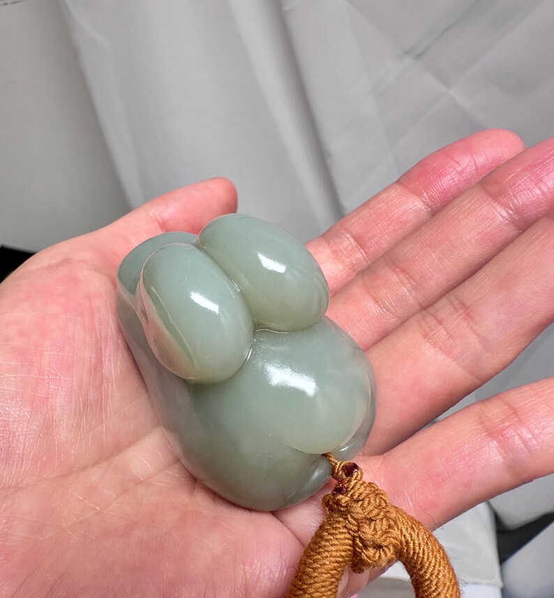 Puede incluir: Un colgante de jade verde p&aacute;lido, liso y de forma org&aacute;nica. Presenta tres protuberancias ovaladas y una base redondeada. Una cuerda trenzada marr&oacute;n est&aacute; unida. El colgante se sostiene en una mano. El colgante de jade mide unos 6 cm de largo.