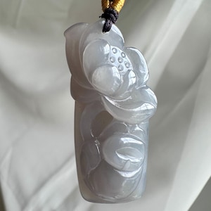 May include: A translucent, pale lavender-gray carved stone pendant featuring a detailed floral motif with a central lotus blossom and rounded petals. The pendant is suspended from a dark brown cord with a small, bright yellow accent knot at the top.