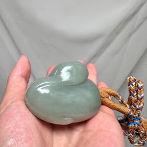 Puede incluir: Una talla de jade verde p&aacute;lido con forma de pato. La piedra pulida tiene un acabado brillante. Un cord&oacute;n decorativo con hilos coloridos est&aacute; unido a la talla.