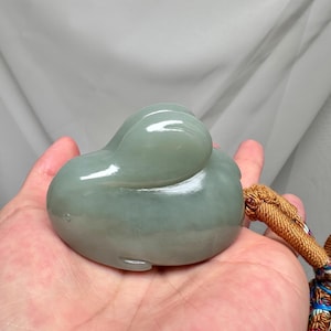 Puede incluir: Una talla de jade verde p&aacute;lido de un animal estilizado, posiblemente un pato o una ballena, sostenida en una mano. La piedra lisa y pulida tiene una forma redondeada con una peque&ntilde;a cola y una cabeza curvada. Una borla marr&oacute;n est&aacute; unida a la talla.