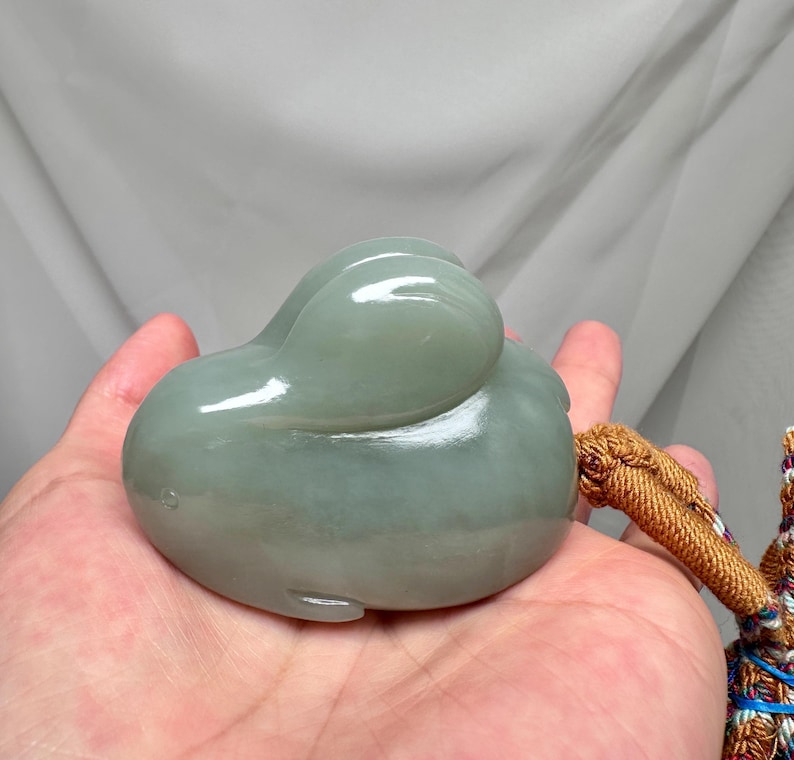 Puede incluir: Una talla de jade verde p&aacute;lido de un animal estilizado, posiblemente un pato o una ballena, sostenida en una mano. La piedra lisa y pulida tiene una forma redondeada con una peque&ntilde;a cola y una cabeza curvada. Una borla marr&oacute;n est&aacute; unida a la talla.