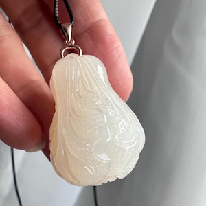 May include: A pear-shaped white translucent stone pendant featuring intricate carved floral and vine patterns. The pendant is suspended from a thin black cord by a polished rose gold-toned metal bail, showcasing a smooth and glossy surface finish.