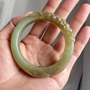 May include: A translucent pale green jade bangle bracelet featuring a carved floral motif along the outer surface. The circular accessory displays natural color variations ranging from light sage to a warmer brownish-yellow hue on one side.