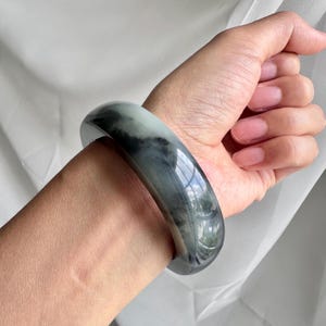 May include: A smooth, polished bangle bracelet crafted from stone with a translucent appearance. The accessory features a marbled pattern with deep charcoal gray and black inclusions set against a pale, milky white and light green base color.