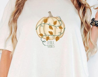May include: Cream-colored t-shirt featuring a pumpkin design with vines and leaves. The pumpkin is surrounded by a vine with leaves and the words "Fall Vibes" in a script font. The shirt is a relaxed fit.