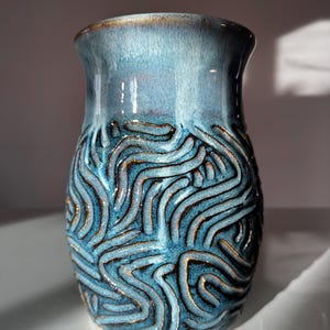 May include: A handcrafted ceramic vase with a glossy, blue glaze. The vase features a textured, wave-like pattern in a darker blue with gold accents. The rim has a subtle, brown hue.