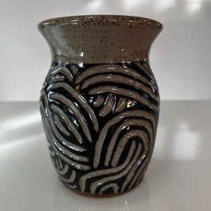 May include: A handmade ceramic vase with a dark brown speckled rim and a black and gray swirling pattern. The vase has a wide opening and a rounded body. The design is a series of curved lines that create a textured effect.