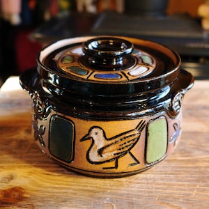 May include: A ceramic casserole dish with a lid. The dish is black with a tan band featuring a bird design. The lid has a decorative pattern with various colors. The dish has handles on either side.