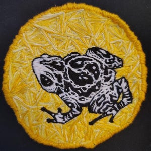 May include: Embroidered patch featuring a black and white frog design on a yellow background. The frog is detailed with intricate patterns, set against a textured, stitched yellow circle. The patch is likely for clothing or accessories.