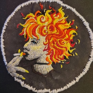 May include: Embroidered patch featuring a woman with fiery orange, yellow, and red hair, smoking a cigarette. The patch has a black background and a white stitched border. The woman's face is detailed with blue eyes and yellow nail polish.