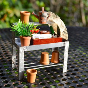 May include: Miniature metal potting bench with a shelf holding a terracotta pot with a green plant, a watering can, and a small straw hat. The bench also has a shelf with three terracotta pots and a small pot on the ground.