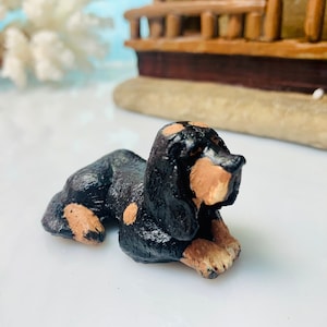 May include: A small, handcrafted figurine of a dachshund dog, lying down. The dog is primarily black with tan markings on its face, paws, and chest. The figurine is set against a blurred background with a miniature ship.
