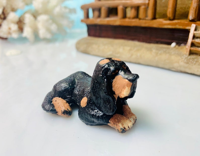 May include: A small, handcrafted figurine of a dachshund dog, lying down. The dog is primarily black with tan markings on its face, paws, and chest. The figurine is set against a blurred background with a miniature ship.