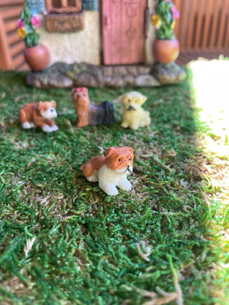 May include: Miniature figurines of dogs in various poses, including a brown and white bulldog, a Yorkshire Terrier, and a yellow puppy, are arranged on a bed of green moss. A small fairy house is in the background, with a pink door and flower pots.