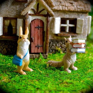 May include: A miniature scene featuring a rabbit with a blue satchel and a squirrel carrying books. The figurines are set in front of a miniature house with a red door and stone facade, on a bed of green moss.