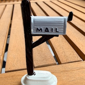 May include: A miniature mailbox figurine with a white base, a dark brown post, and a silver mailbox. The word "MAIL" is printed on the front of the mailbox. The figurine is made of ceramic or a similar material.
