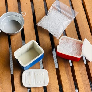 May include: A collection of miniature dollhouse accessories, including a small silver bucket, a blue and white cooler, a red and white cooler, a clear plastic bag filled with small clear beads, and a white lid. The items are arranged on a wooden surface.