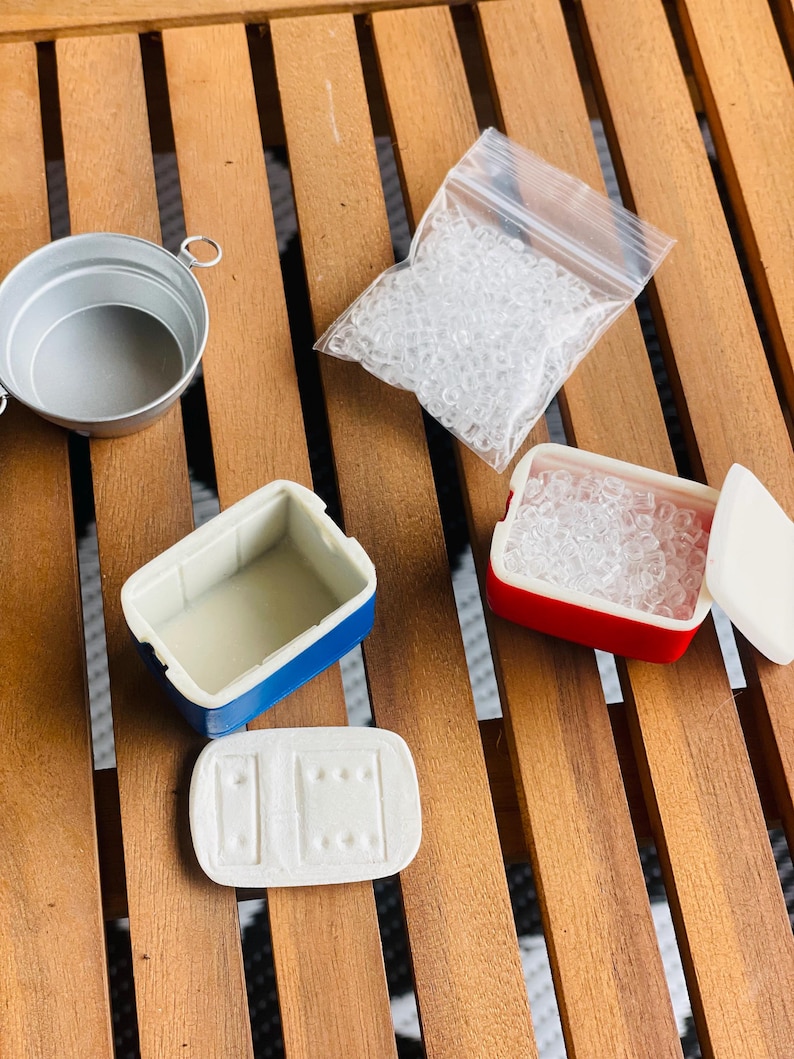 May include: A collection of miniature dollhouse accessories, including a small silver bucket, a blue and white cooler, a red and white cooler, a clear plastic bag filled with small clear beads, and a white lid. The items are arranged on a wooden surface.