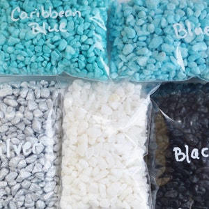 May include: Assortment of small decorative stones in clear plastic bags. Colors include Caribbean Blue, Blue, Silver, White, and Black. Each bag is labeled with the color name in handwritten script. These stones are likely used for crafts or decorative purposes.