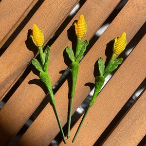 May include: Three miniature corn stalks with yellow corn and green leaves. The corn is a bright yellow color, and the leaves are a vibrant green. The stalks are arranged on a wooden surface.