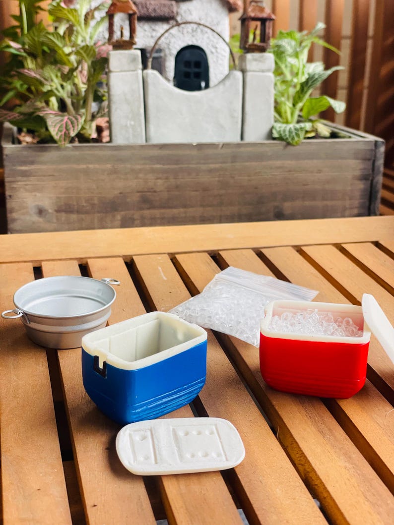 May include: Miniature picnic set with a blue and white cooler, a red and white cooler, a small metal bucket, and a bag of clear ice. The set is on a wooden surface, with a miniature garden in the background.