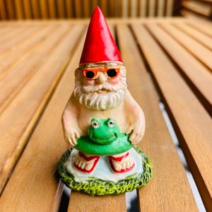 May include: A whimsical garden gnome figurine with a red pointed hat and orange sunglasses. The gnome is holding a green frog-shaped floatie and wearing red and white sandals. The figurine is on a green and white base.