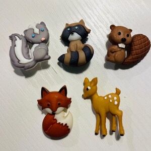 May include: A collection of five animal-shaped decorative items. The set includes a skunk, raccoon, beaver, fox, and deer. The skunk is gray and white, the raccoon is brown and black, the beaver is brown, the fox is red and white, and the deer is yellow with white spots.