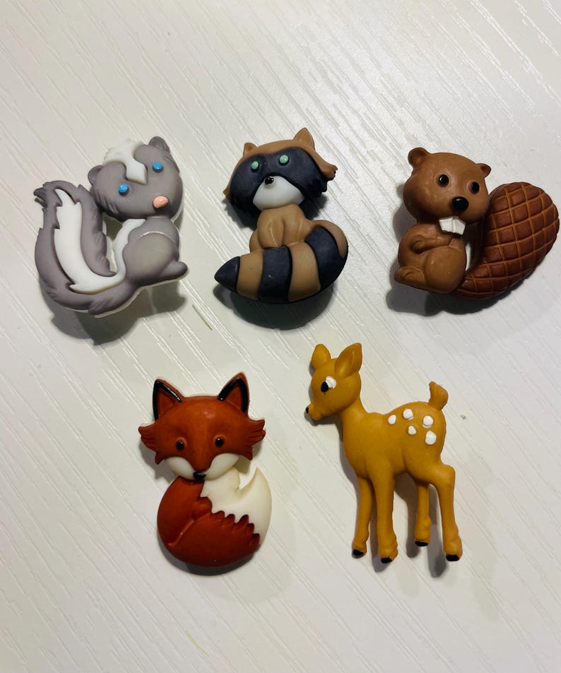 May include: A collection of five animal-shaped decorative items. The set includes a skunk, raccoon, beaver, fox, and deer. The skunk is gray and white, the raccoon is brown and black, the beaver is brown, the fox is red and white, and the deer is yellow with white spots.