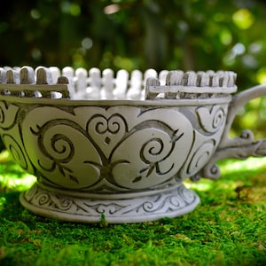 May include: A decorative, off-white teacup with a detailed, carved design. The cup features a handle and a rim with a fence-like structure. The cup is placed on a bed of green moss, with a blurred green background.