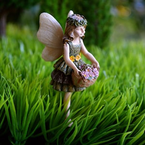 May include: A miniature fairy figurine with delicate wings and a floral crown, holding a basket of purple flowers. The fairy is dressed in a green and brown layered dress. The figurine is set in a bed of vibrant green grass.