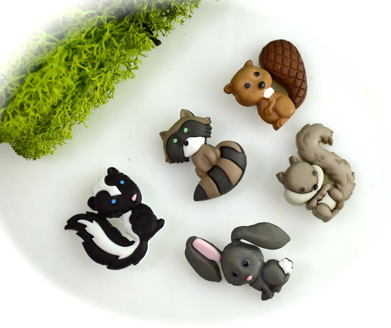 May include: A collection of whimsical woodland animal figurines, including a skunk, raccoon, beaver, squirrel, and rabbit. The figurines are in various shades of brown, gray, black, and white, with details like striped tails and pink ears. They are arranged on a white surface.