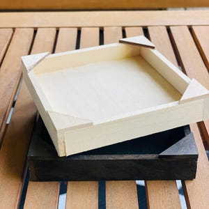 May include: Two wooden trays, one light-colored and one dark, are stacked on a wooden surface. The light-colored tray is square with corner supports. The dark tray is also square and sits beneath the light-colored tray.