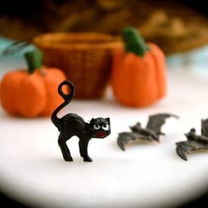 May include: A Halloween-themed still life featuring a black cat figurine with a curled tail, orange pumpkins with green stems, and gray bat figurines. A small wicker basket is in the background.