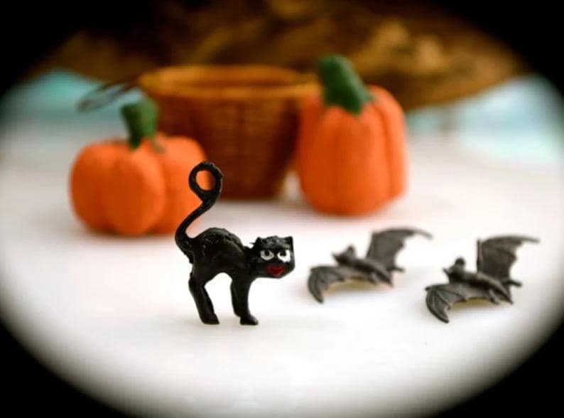 May include: A Halloween-themed still life featuring a black cat figurine with a curled tail, orange pumpkins with green stems, and gray bat figurines. A small wicker basket is in the background.