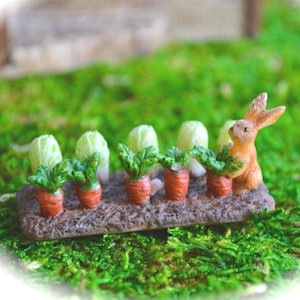 May include: A miniature figurine set featuring a brown rabbit standing next to a row of orange carrots with green leafy tops and pale green cabbages, all arranged on a textured brown rectangular base representing a garden plot of soil.
