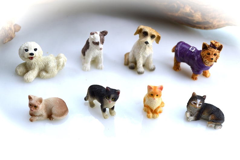 May include: A collection of miniature figurines of dogs and cats. The dogs include a white poodle, a brown and white dog, a tan dog, and a Yorkshire Terrier in a purple coat. The cats are in various poses and colors.