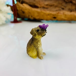 May include: A miniature figurine of a seated dog with a purple flower on its head. The dog has a light brown coat and black eyes. The figurine is likely made of resin or a similar material.