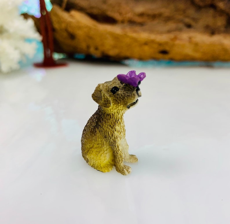 May include: A miniature figurine of a seated dog with a purple flower on its head. The dog has a light brown coat and black eyes. The figurine is likely made of resin or a similar material.