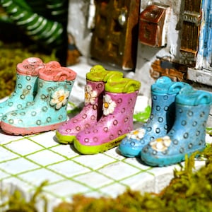 May include: Miniature colorful rain boots in shades of blue, pink, and purple, adorned with white flower details and polka dots. The boots are arranged on a white tiled surface, suggesting a garden or outdoor theme.