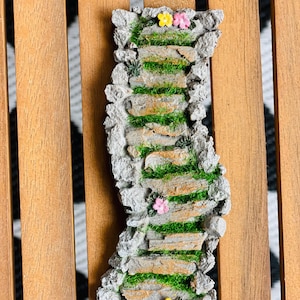 May include: A miniature stone staircase with steps of tan-colored stone and patches of green grass. The edges are lined with gray stones. Small yellow and pink flowers are placed at the top and bottom of the staircase.