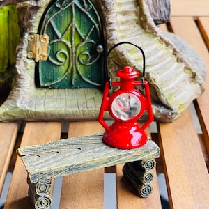 May include: A miniature red lantern with a black handle sits on a small wooden bench. The bench has a weathered appearance, with a textured surface and log-like supports. In the background is a green door with ornate detailing, part of a fairy house.