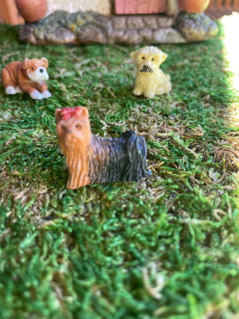 May include: Miniature figurines of three different dog breeds on a bed of green moss. The dogs are a bulldog, a terrier, and a small, fluffy dog. The bulldog is brown and white, the terrier is black and tan, and the fluffy dog is yellow.