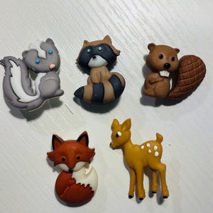 May include: A collection of five animal-shaped decorative embellishments. The set includes a skunk, raccoon, beaver, fox, and deer. The skunk is gray and white, the raccoon is brown and black, the beaver is brown, the fox is red and white, and the deer is yellow with white spots.