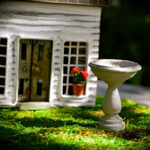 May include: A miniature white house with a detailed facade, featuring windows and a door. A small bird bath stands in front of the house, set on a bed of green moss. Red flowers are visible in a window.